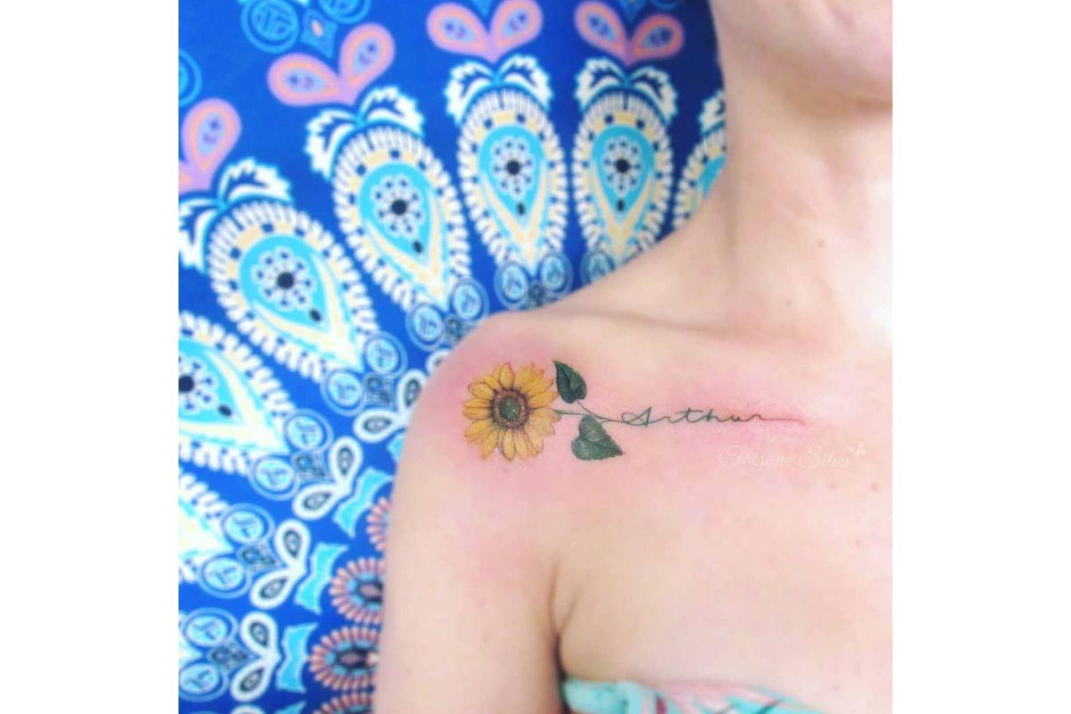 Tatuagem com nome de filho no ombro usando letra cursiva e com desenho de flor