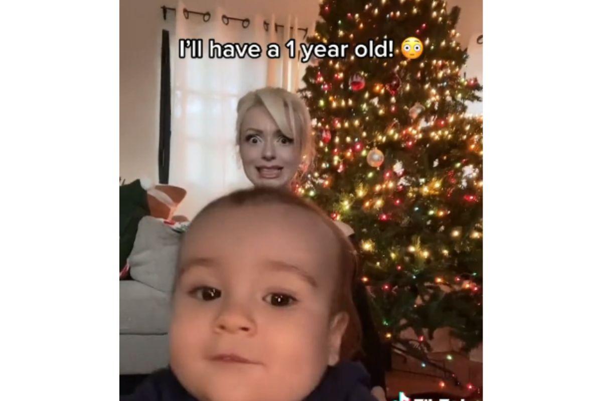 Mulher branca com cabelo loiro com filho no colo e com filho no colo atrás de árvore de natal