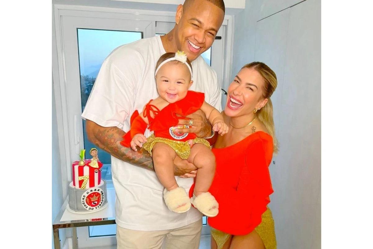 Mulher branca com cabelo loiro e blusa vermelha, homem com blusa branca com filha no colo