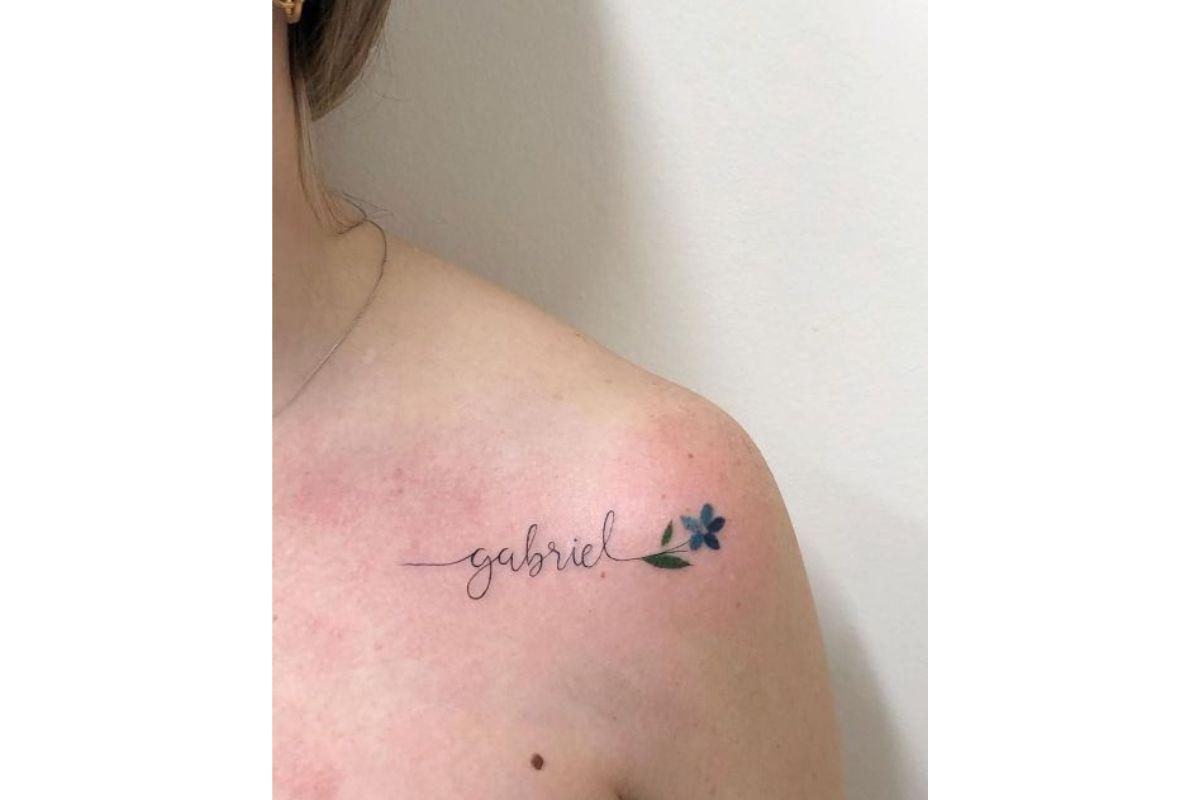 Tatuagem com nome de filho no ombro usando letra cursiva e com desenho de flor