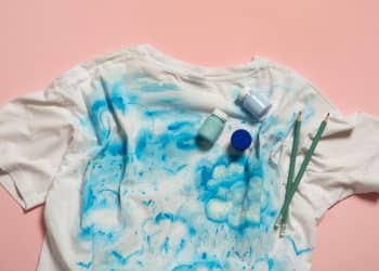 Camiseta branca com manchas azuis em fundo rosa, representando roupas sujas de crianças.