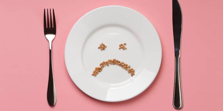 Prato com grãos formando uma carinha triste, acompanhado por garfo e faca em um fundo rosa, simbolizando a alergia alimentar.