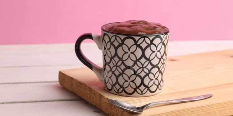 Brownie em uma caneca em cima de uma tábua de madeira com fundo rosa.