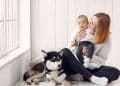 Mãe, bebê e pet em um ambiente de cor clara com janela.