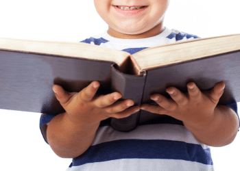 Criança com um livro grande na mãe e sorrindo. Na imagem, ela está em um fundo branco