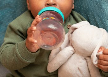 Um bebê segurando uma mamadeira de água e agarrrado em um ursinho