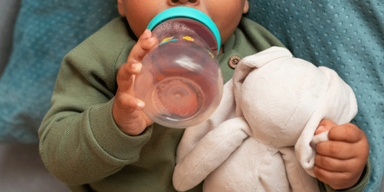 Um bebê segurando uma mamadeira de água e agarrrado em um ursinho