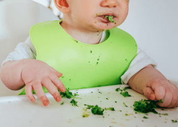Criança comendo, provavelmente um=algum alimento que seja folha. Ela está com um babador verde e com a boca suja
