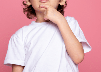 Criança com a mão ma boca, uma camisa branca e um fundo rosa.