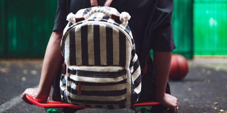 Jovem sentado num skate com uma mochila maluca nas costas.