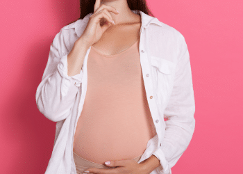 Mulher grávida com a mão na barriga, a imagem está em um fundo rosa.