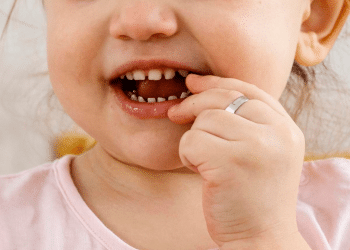 Criança com dedo na boca, dentes aparentes e camiseta rosa