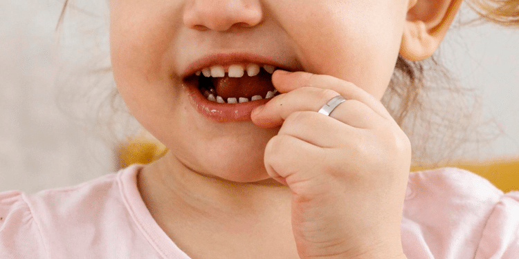 Criança com dedo na boca, dentes aparentes e camiseta rosa