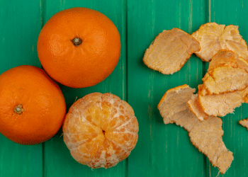 Três tangerinas sob fundo verde, de madeira. Duas estão com casca e uma encontra-se descascada, com suas cascas ao lado.