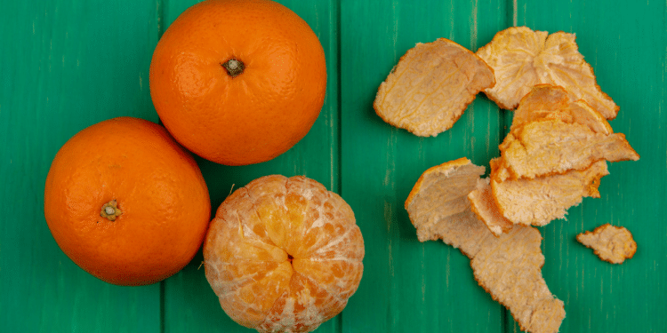 Três tangerinas sob fundo verde, de madeira. Duas estão com casca e uma encontra-se descascada, com suas cascas ao lado.