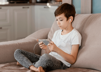 Criança sentada no sofá, usando fones de ouvido e mexendo no celular