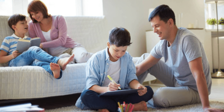Familia reunida na sala enquanto pai ensina o dever para o menino mais velho, a mae ri e brinca com mais novo no sofá